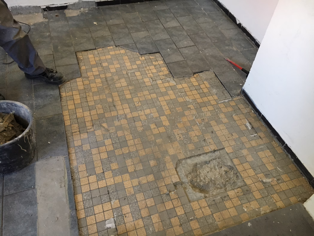 UZIN_Application_Image_UZIN NC 110_0445_ 2018-03 Delvis fjernet flisgulv under renovering: i forgrunnen ligger små beige‑grå mosaikkfliser med slitte og manglende partier; rundt kanten er større mørke rektangulære fliser løftet, og lim og undergulv er synlig. Til venstre sees en arbeiders bein og svarte støvler ved siden av en bøtte med fjernet flis; til høyre lener en rød brekkstang mot veggen. Hvite malte vegger med svart gulvlist, støv og rusk på gulvet. Bildet er tatt fra lav vinkel nedover.